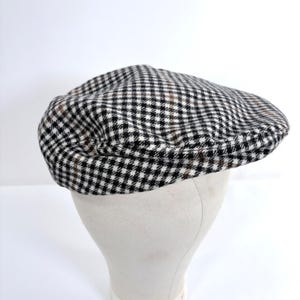 May include: A black, white, and brown checkered flat cap. The hat is made of a textured fabric and is displayed on a white mannequin head. The cap has a rounded crown and a small, stiff brim. The pattern is a classic houndstooth design.