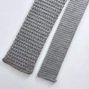 May include: Two grey neckties displayed side by side. The left tie features a coarse, textured knit pattern with a visible weave, while the right tie has a finer, horizontal ribbed knit construction. Both items are long, narrow, and rectangular.