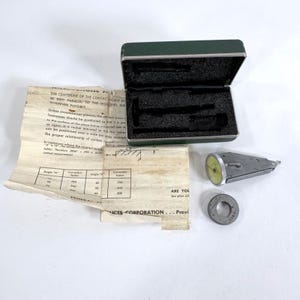 May include: An open green metal case with black foam inserts, a silver measuring tool with a yellow dial, a metal ring, and a folded paper with text and diagrams. The text on the paper includes "THE CENTERLINE OF THE CONTACT POINT".