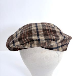 May include: A brown and beige plaid newsboy cap. The cap features a classic plaid pattern with dark blue and red accents. The hat is displayed on a white mannequin head against a white background.