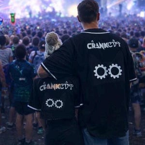 May include: Two people wearing black baseball-style shirts with white trim. The shirts have the word "CRANKED" arched across the top and two white gear icons below. The background is a crowded outdoor event.