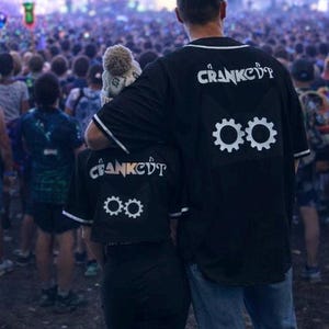 May include: Two people wearing black baseball-style shirts with the word "CRANKCDT" in white script on the back. Below the text are two white gear symbols. The shirts have white trim and are worn at a concert.