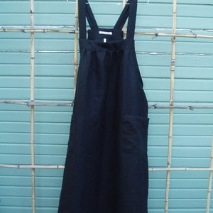 May include: A black linen apron with adjustable straps and a single pocket. The apron is hanging on a silver hanger against a light blue background.