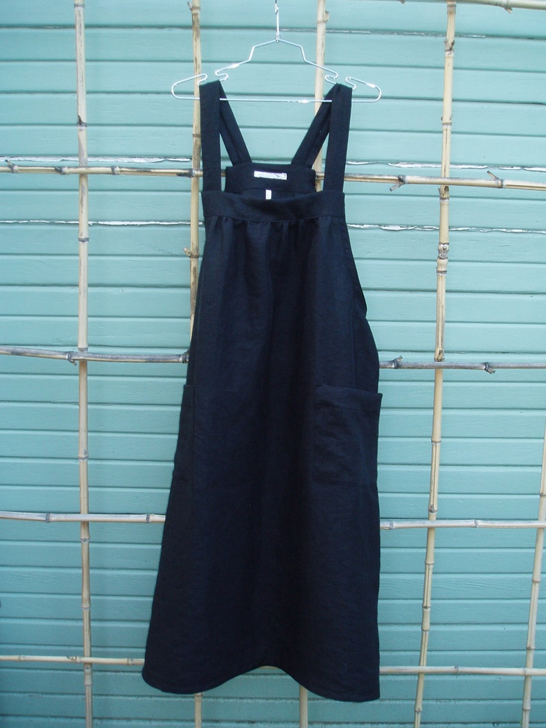 May include: A black linen apron with adjustable straps and a single pocket. The apron is hanging on a silver hanger against a light blue background.