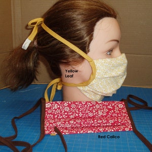 May include: Two fabric face masks. One is yellow with a leaf pattern, the other is red with a white floral design. Both have tie closures. The yellow mask is on a mannequin head. The red mask is labeled "Red Calico".