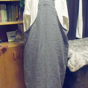 May include: A gray linen apron with adjustable straps. The apron is hanging on a white hanger and has a white ruffle at the bottom.