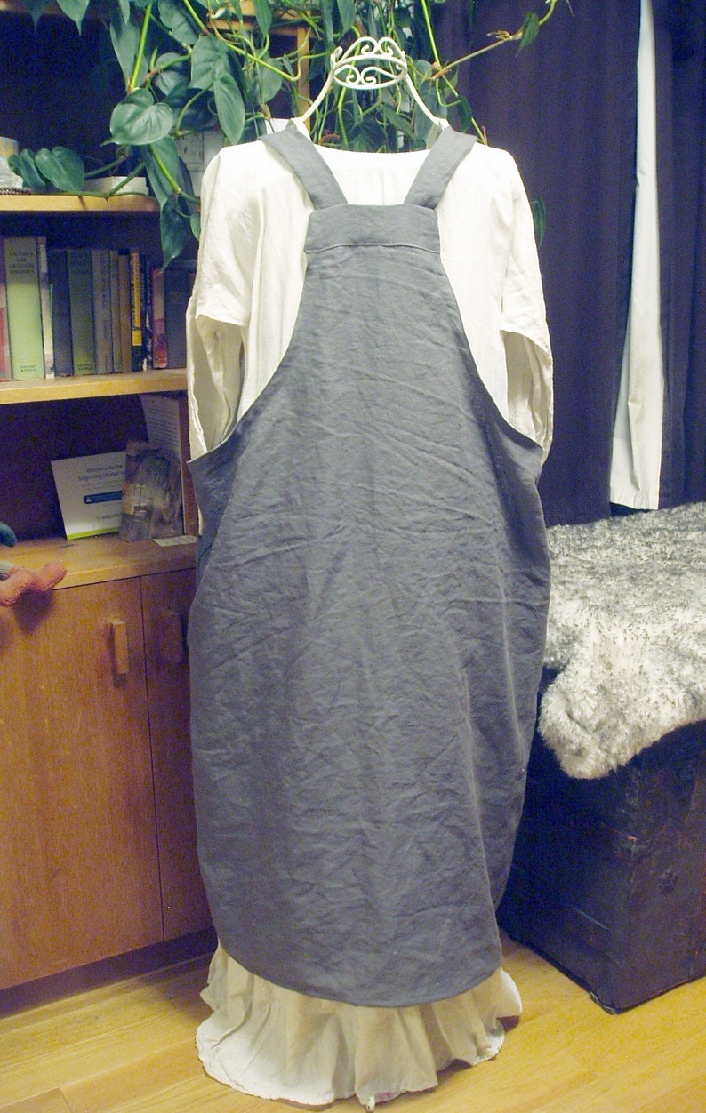 May include: A gray linen apron with adjustable straps. The apron is hanging on a white hanger and has a white ruffle at the bottom.