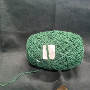 May include: A ball of dark green yarn with a textured, open-weave appearance. A small, rectangular white paper label is attached to the center of the yarn ball, featuring printed text and numbers that are partially obscured and difficult to read.