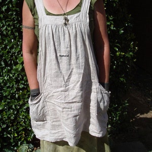 May include: A beige linen apron with a green top and a brown necklace. The apron has a large pocket on the front and the word "Natural" is printed on the front.