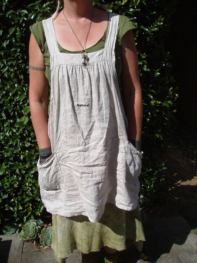 May include: A beige linen apron with a green top and a brown necklace. The apron has a large pocket on the front and the word "Natural" is printed on the front.