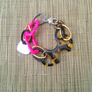 Puede incluir: Una pulsera multicolor con varios elementos. Incluye una cadena rosa brillante, un dije de perro con estampado de carey y eslabones metálicos en plata, dorado y negro. También tiene una pequeña etiqueta plateada.
