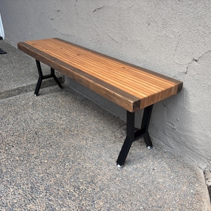 Può includere: Panca in legno con finitura naturale e gambe in metallo nero. La panca ha un piano a doghe ed è lunga circa 152 cm. Le gambe sono angolate per stabilità. La panca è posizionata su una superficie di cemento accanto a un muro grigio.