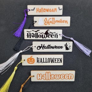 Puede incluir: Marcapáginas de madera con temática de Halloween. Cada marcapáginas presenta la palabra 'Halloween' en naranja o negro, junto con ilustraciones espeluznantes. Los marcapáginas están acabados con borlas de colores.