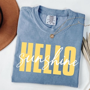 Può includere: Una t-shirt blu a maniche corte con scollo rotondo ed etichetta del marchio Comfort Colors. La maglietta presenta la parola HELLO in grandi lettere gialle e la parola sunshine scritta in corsivo bianco sovrapposta al testo.