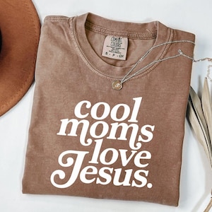 May include: A brown t-shirt with the text "cool moms love Jesus" in white script. The shirt has a crew neck and short sleeves. A silver necklace with a small pendant is draped across the shirt.