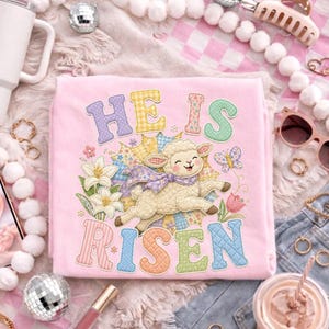 May include: A pink t-shirt with the words "HE IS RISEN" in colorful letters. The shirt features an embroidered lamb, flowers, and a butterfly. Other items include a white tumbler, sunglasses, and a phone with a heart image.