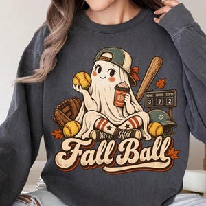 May include: A charcoal grey crewneck sweatshirt featuring a graphic of a ghost wearing a baseball cap, holding a softball and a coffee cup. The design includes a baseball bat, glove, scoreboard reading 3-7-2, and the text Fall Ball in script.