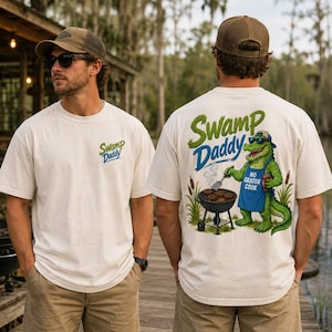 May include: Off-white t-shirts with the text "Swamp Daddy" in green and blue. One shirt has the text on the front, and the other has a cartoon alligator grilling burgers on the back, with the text "No Gator Cook".