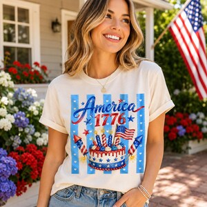 May include: Cream-colored t-shirt with a patriotic design. The shirt features the word "America" in blue script, the year "1776", and a cake with candles, fireworks, and an American flag. The design is set against blue and white stripes.