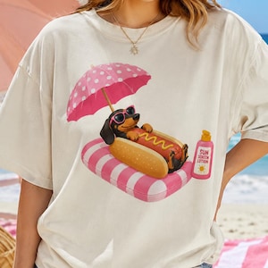 May include: Cream-colored t-shirt featuring a cartoon dachshund wearing sunglasses, lounging on a pink and white striped float, under a pink polka-dot umbrella. A hot dog and sunscreen bottle are also depicted.
