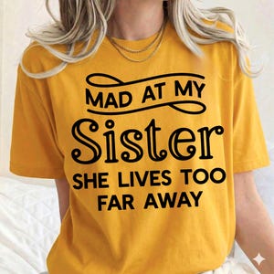 May include: Mustard yellow t-shirt with the text "MAD AT MY Sister SHE LIVES TOO FAR AWAY" in black. The shirt has short sleeves and a crew neck. The text is in a bold, playful font.