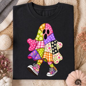 Puede incluir: Una camiseta negra con un gráfico central de un fantasma hecho de varios patrones de tela de patchwork coloridos, incluyendo estampados florales, de lunares y de cuadros. El fantasma lleva zapatillas estampadas rosas y moradas con cordones.