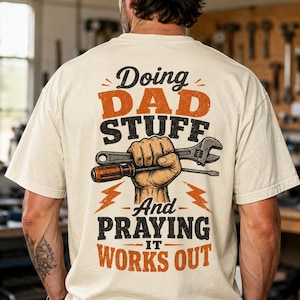 May include: Cream-colored t-shirt with the text "Doing Dad Stuff" in black and orange, and "And Praying It Works Out" in black and orange. A hand graphic holds a wrench and screwdriver. The shirt is worn by a person.