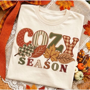 Może przedstawiać: Kremowy t-shirt z napisem "COZY SEASON" wykonanym dużymi, ozdobnymi literami. Każda litera oraz towarzyszące jej grafiki liści jesiennych wypełnione są różnymi wzorami, w tym kropkami, paskami i kratką w odcieniach pomarańczu, brązu i zieleni.