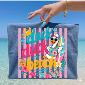 May include: Blue t-shirt with a colorful graphic featuring a cartoon duck wearing sunglasses, a Hawaiian shirt, and holding a surfboard. The text "duck duck beach" is displayed in vibrant colors, with pink and white stripes in the background.