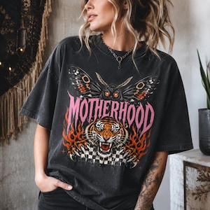 Op de afbeelding: Een antracietgrijs t-shirt met korte mouwen en een grafische print van een mot boven een tijgerkop, omringd door oranje vlammen. Het woord MOTHERHOOD staat in grote roze letters in het midden gedrukt, boven een zwart-wit geblokt patroon.