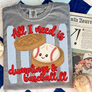 Könnte beinhalten: Graues Comfort Colors T-Shirt mit dem Text "All I need is dumplings & baseball". Die Grafik zeigt eine als Baseball gestaltete Teigtasche. Das Shirt ist ein bequemer, lässiger Stil.