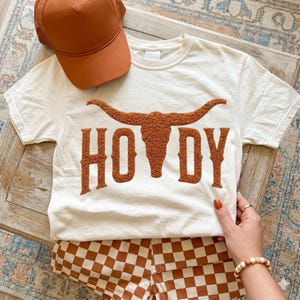 May include: Cream-colored t-shirt with the word "HOWDY" in brown, featuring a longhorn graphic. Paired with brown and white checkered shorts and a brown trucker hat. A casual, western-themed outfit.