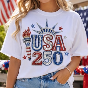 Könnte beinhalten: Weißes T-Shirt mit einem patriotischen Design mit dem Text "USA 250" in roten, weißen und blauen Strasssteinen. Das Design umfasst eine Fackel, Sterne und eine Krone, die den 250. Jahrestag der Vereinigten Staaten feiert.