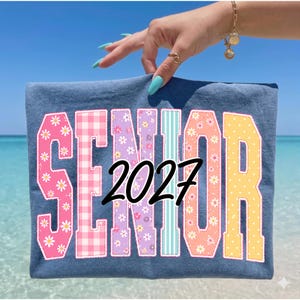 以下が含まれることがあります： カラフルな花柄とチェック柄の文字で「SENIOR」と黒で「2027」と書かれた青いTシャツ。青い空と海の背景にシャツが表示されています。