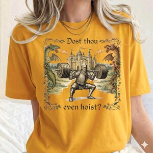 May include: Mustard yellow t-shirt featuring a medieval knight lifting weights in front of a castle. The design includes dragons and the text "Dost thou even hoist?" in a decorative frame.