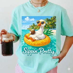 Puede incluir: Camiseta azul claro con un diseño de un ganso con gafas de sol flotando en un flotador con forma de piña en el océano, sosteniendo una bebida de coco. El texto "Sippin' Pretty" está debajo, con un fondo tropical.