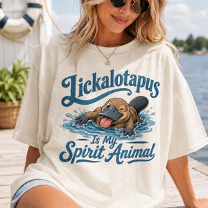 Può includere: T-shirt color crema con un disegno di ornitorinco nell'acqua e la scritta blu "Tickalotapus Is My Spirit Animal". L'ornitorinco è marrone con la lingua rosa e fa l'occhiolino. La maglietta ha una vestibilità ampia.