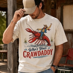 Puede incluir: Camiseta color crema con un gráfico de un vaquero montando un gran cangrejo rojo. El texto "Who's Your Crawdaddy" está impreso debajo en rojo y azul. Es una prenda de vestir informal.