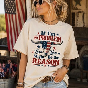 May include: Cream-colored t-shirt with the text "If I'm the PROBLEM Then You Might Be the REASON" and a bull skull design with an American flag pattern. The shirt is paired with denim shorts and a brown belt. An American flag is in the background.