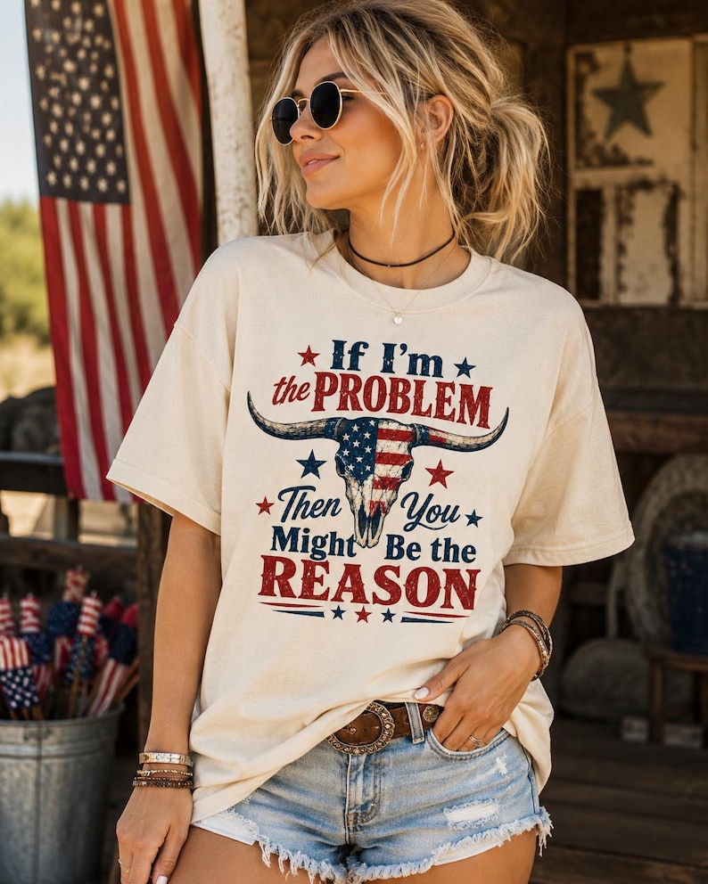 May include: Cream-colored t-shirt with the text "If I'm the PROBLEM Then You Might Be the REASON" and a bull skull design with an American flag pattern. The shirt is paired with denim shorts and a brown belt. An American flag is in the background.