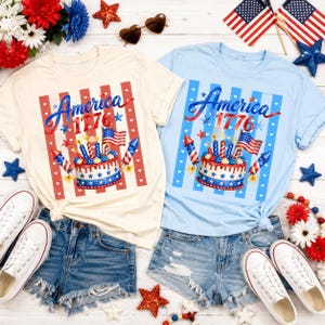 May include: Two t-shirts, one cream and one light blue, with a patriotic design. The design includes the word "America 1776" and a cake with candles, flags, and fireworks. Paired with denim shorts and white sneakers.