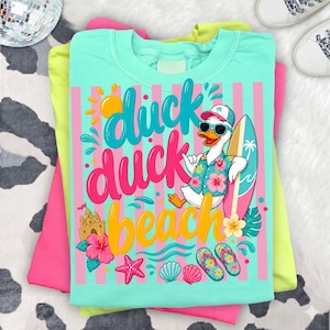May include: A stack of t-shirts in pink, yellow, and turquoise. The top shirt features a graphic of a duck wearing sunglasses and a Hawaiian shirt, holding a surfboard. The text "duck duck beach" is displayed in colorful lettering.