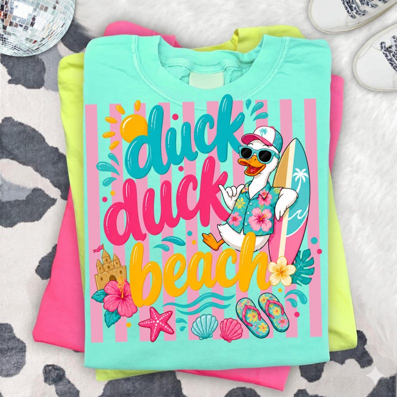 May include: A stack of t-shirts in pink, yellow, and turquoise. The top shirt features a graphic of a duck wearing sunglasses and a Hawaiian shirt, holding a surfboard. The text "duck duck beach" is displayed in colorful lettering.