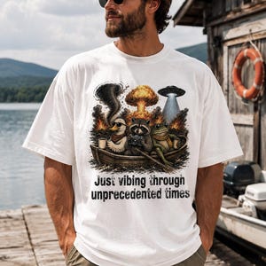 May include: A white short-sleeve t-shirt featuring a graphic of a duck, a raccoon, and a frog in a wooden boat. The scene includes a tornado, an explosion, and a UFO. The text below the graphic reads, "Just vibing through unprecedented times."