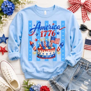 May include: Light blue sweatshirt with a patriotic design. The graphic features the word "America 1776" in red and blue script, along with a cake, fireworks, and an American flag. The sweatshirt is a long-sleeved crew neck.