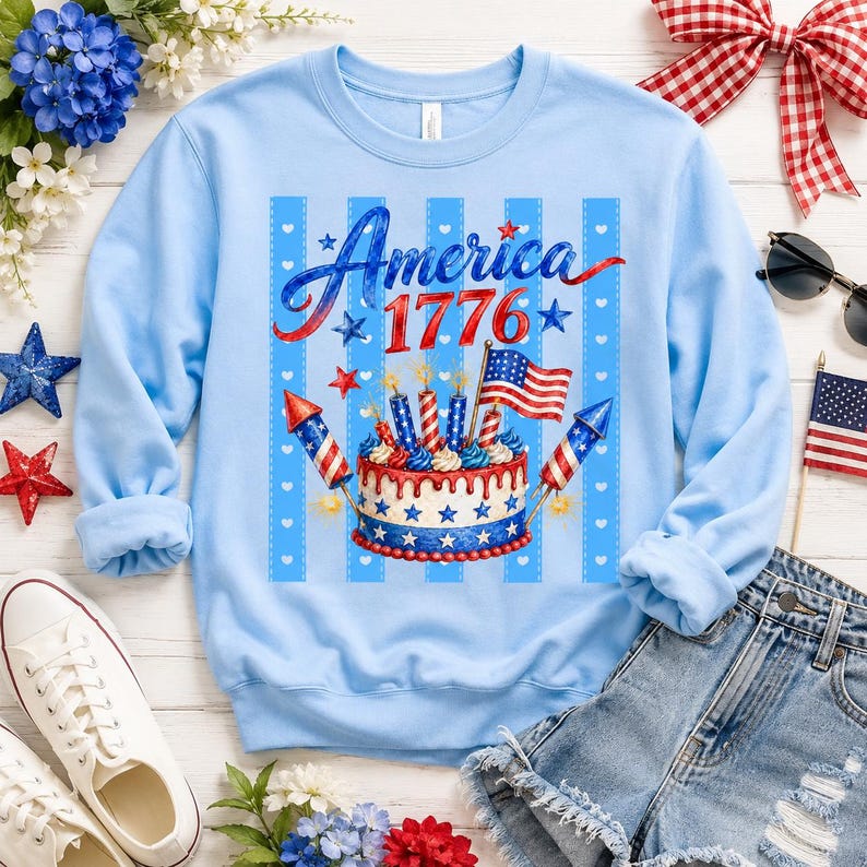 May include: Light blue sweatshirt with a patriotic design. The graphic features the word "America 1776" in red and blue script, along with a cake, fireworks, and an American flag. The sweatshirt is a long-sleeved crew neck.