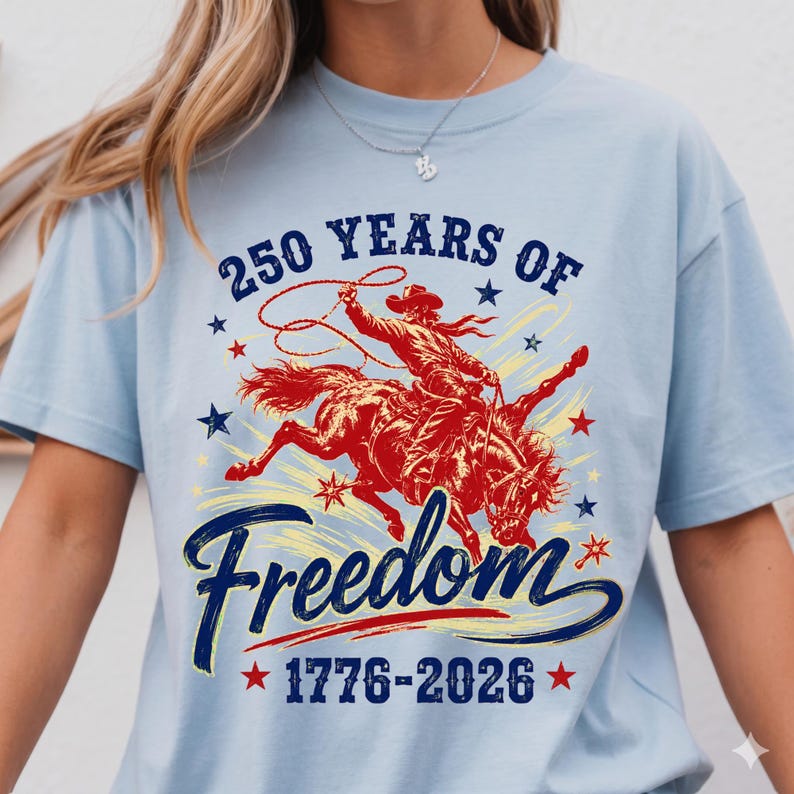 May include: Light blue t-shirt featuring a graphic of a cowboy riding a bucking bronco, with the text "250 Years of Freedom 1776-2026". The design is in red and navy blue, with star accents.