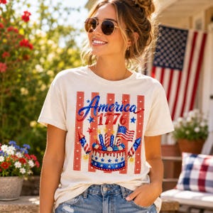 May include: Cream-colored t-shirt with a patriotic design. The graphic features the word "America" in blue script, the year "1776", a cake with candles, an American flag, and red and blue stripes. The shirt is worn by a person.