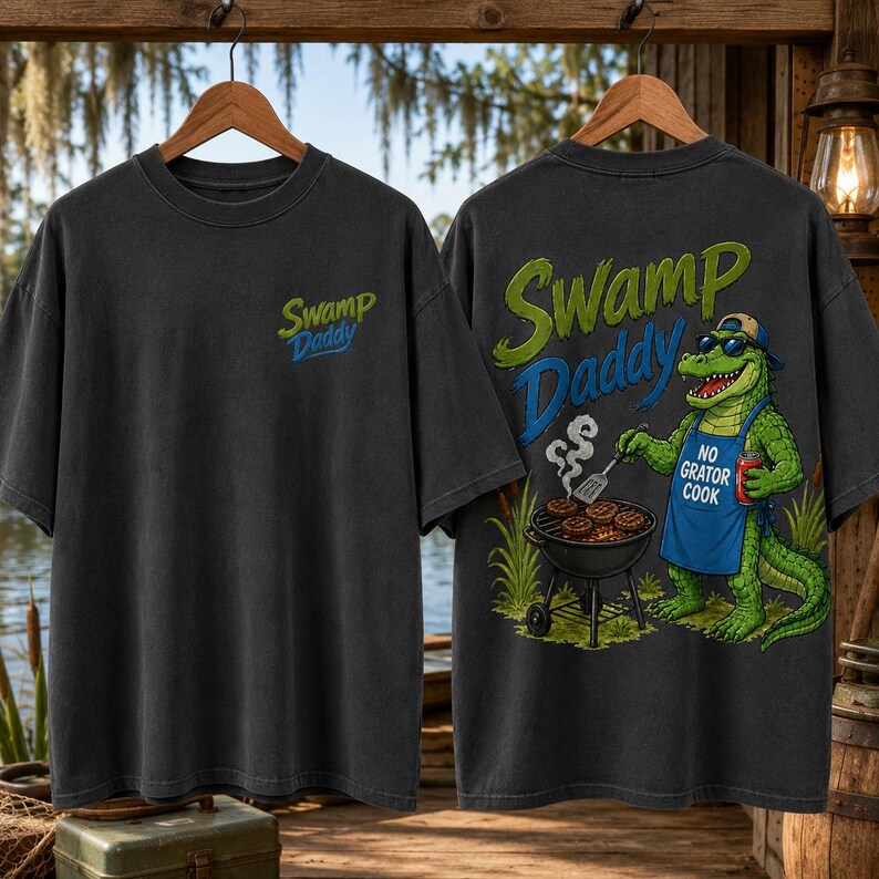 May include: Dark gray t-shirts with the text "Swamp Daddy" in green and blue. One shirt has the text on the front. The other shirt has a cartoon alligator grilling burgers with the text "No Gator Cook".