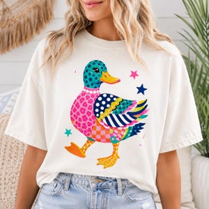 May include: An off-white short-sleeve t-shirt featuring a colorful graphic of a duck. The duck has a teal head with polka dots, a pink floral patterned chest, and a wing with blue, yellow, and white stripes. Small pink and blue stars surround it.